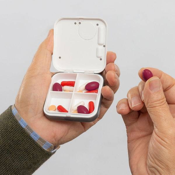 Persoană ținând o pastilă și cutia inteligentă Pilly cu 4 compartimente, ideală pentru dozarea corectă a medicamentelor în rutina zilnică.